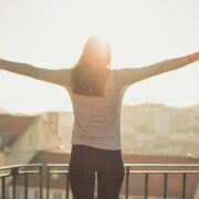 A woman standing on a balcony with arms outstretched, embracing the morning sunlight, symbolizing empowerment, personal growth, and the freedom achieved through life coaching.