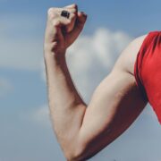 A close-up of a certified health coach's flexed arm, showcasing toned muscles. The person is wearing a red sleeveless shirt and a black ring on their finger. The background features a blue sky with white clouds.