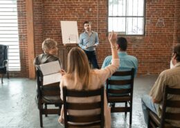 Group coaching session with a diverse group of adults, one participant raising her hand while the coach leads a discussion in a brick-walled room.