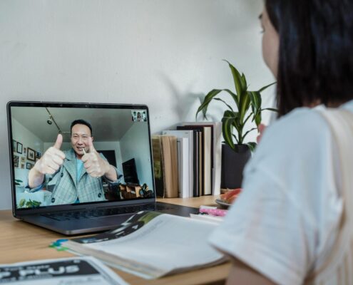 A coach and client communicate through a video call on a laptop. The coach on the screen is smiling and giving two thumbs up, while the client sits at a desk with notebooks, a laptop, and books nearby, symbolizing an online coaching session and positive connection.
