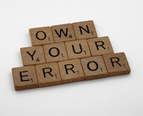 Scrabble tiles spelling “Own Your Error” on a wooden surface, symbolizing personal responsibility, growth, and the power of accountability in coaching.