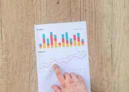 A hand points to a printed financial report showing colorful bar and line graphs representing company performance metrics like sales, receipts, and orders—symbolizing the measurable ROI of life coaching and personal growth.