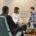 A couple sits on a beige couch during a relationship coaching session, attentively speaking with a coach seated across from them taking notes in a modern, warmly lit office.