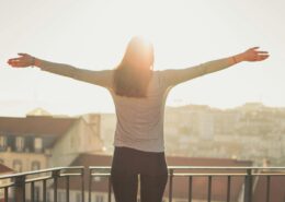 A woman stands on a balcony with her arms outstretched, facing the morning sun over a city skyline. The warm sunlight creates a glowing, hopeful atmosphere, symbolizing confidence, freedom, and self-empowerment — aligning with the theme of overcoming imposter syndrome and embracing authenticity.