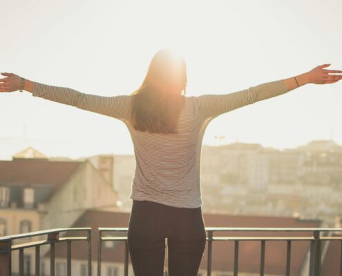 A woman stands on a balcony with her arms outstretched, facing the morning sun over a city skyline. The warm sunlight creates a glowing, hopeful atmosphere, symbolizing confidence, freedom, and self-empowerment — aligning with the theme of overcoming imposter syndrome and embracing authenticity.