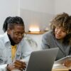 Two people sitting together at a desk having a focused coaching conversation. One person is using a laptop while the other holds a tablet and offers guidance, both smiling and engaged in a collaborative discussion.