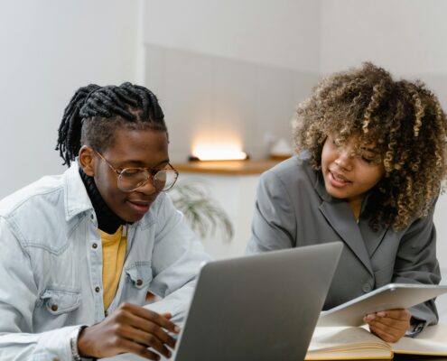 Two people sitting together at a desk having a focused coaching conversation. One person is using a laptop while the other holds a tablet and offers guidance, both smiling and engaged in a collaborative discussion.