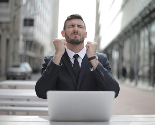 A determined man in a business suit sitting at a laptop outdoors, clenching his fists in a moment of emotional resilience or triumph, symbolizing overcoming setbacks through inner strength.