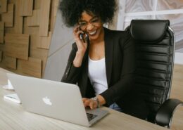Smiling woman sitting in front of a laptop, talking on her phone, celebrating progress—symbolizing how to help a stuck client move forward.