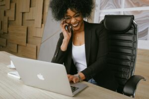 Smiling woman sitting in front of a laptop, talking on her phone, celebrating progress—symbolizing how to help a stuck client move forward.