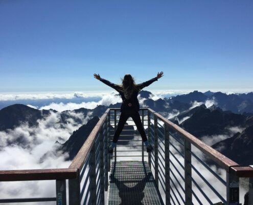 Person standing with arms wide open on a mountain viewpoint above the clouds, symbolizing freedom, possibility, and The Power of AND™.