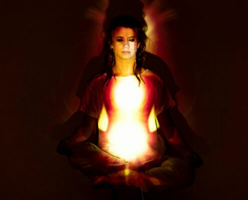 A person sitting cross-legged in a meditative posture with eyes closed, illuminated by warm red and yellow light patterns that create a glowing, hypnotic effect across their chest and face.
