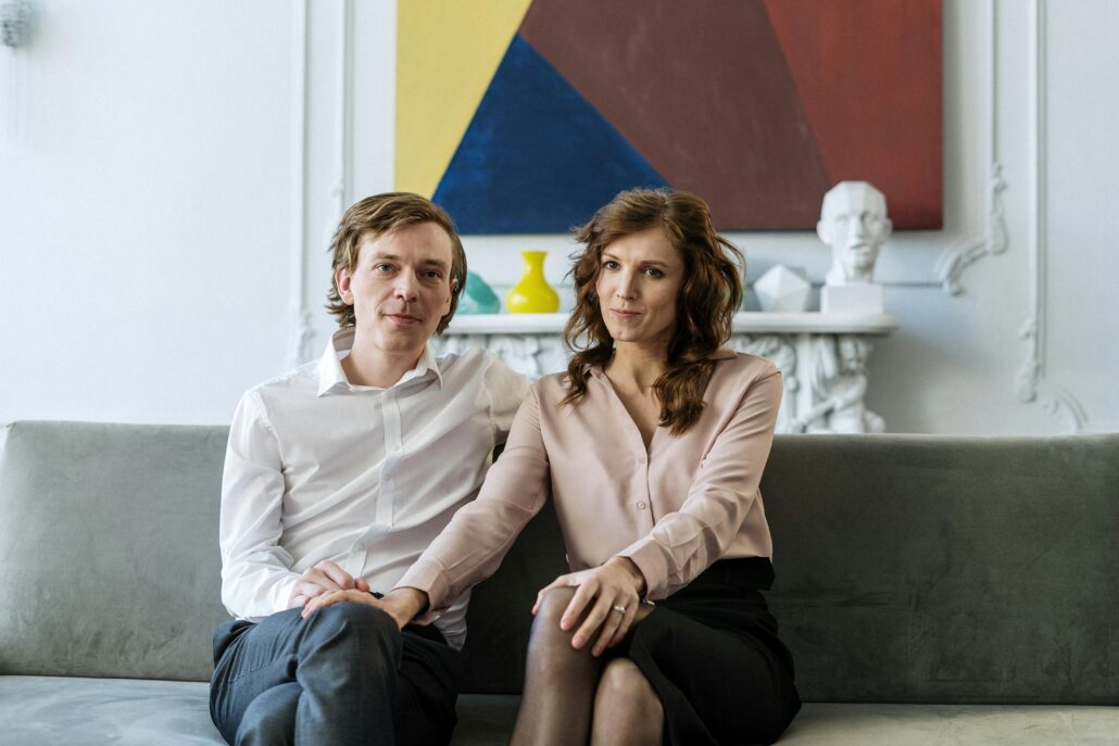 A well-dressed couple sitting close together on a sofa in a modern living room, looking calm and connected—illustrating couples coaching vs couples therapy and the different approaches to strengthening relationships.