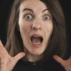 A surprised woman with wide eyes and open mouth holds her hands near her face against a dark background, expressing shock and anxiety—illustrating coaching fear of failure and the emotional overwhelm that can come with self-doubt.