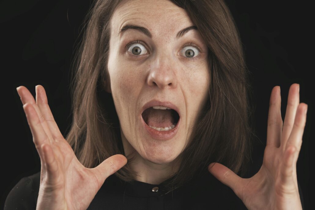 A surprised woman with wide eyes and open mouth holds her hands near her face against a dark background, expressing shock and anxiety—illustrating coaching fear of failure and the emotional overwhelm that can come with self-doubt.