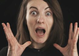 A surprised woman with wide eyes and open mouth holds her hands near her face against a dark background, expressing shock and anxiety—illustrating coaching fear of failure and the emotional overwhelm that can come with self-doubt.