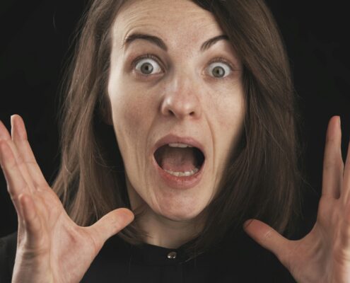 A surprised woman with wide eyes and open mouth holds her hands near her face against a dark background, expressing shock and anxiety—illustrating coaching fear of failure and the emotional overwhelm that can come with self-doubt.