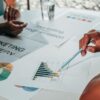 Hands pointing at marketing charts and strategy documents on a table during a collaborative planning session, illustrating a coaching lead magnet focused on attracting and engaging ideal clients.