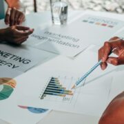 Hands pointing at marketing charts and strategy documents on a table during a collaborative planning session, illustrating a coaching lead magnet focused on attracting and engaging ideal clients.