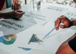 Hands pointing at marketing charts and strategy documents on a table during a collaborative planning session, illustrating a coaching lead magnet focused on attracting and engaging ideal clients.