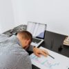 Person experiencing coaching burnout asleep at a desk beside a laptop, financial reports, and work materials, illustrating exhaustion, stress, and overwhelm in a professional coaching environment.
