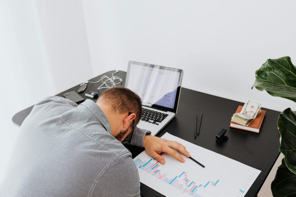 Person experiencing coaching burnout asleep at a desk beside a laptop, financial reports, and work materials, illustrating exhaustion, stress, and overwhelm in a professional coaching environment.