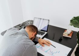 Person experiencing coaching burnout asleep at a desk beside a laptop, financial reports, and work materials, illustrating exhaustion, stress, and overwhelm in a professional coaching environment.