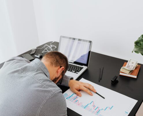 Person experiencing coaching burnout asleep at a desk beside a laptop, financial reports, and work materials, illustrating exhaustion, stress, and overwhelm in a professional coaching environment.