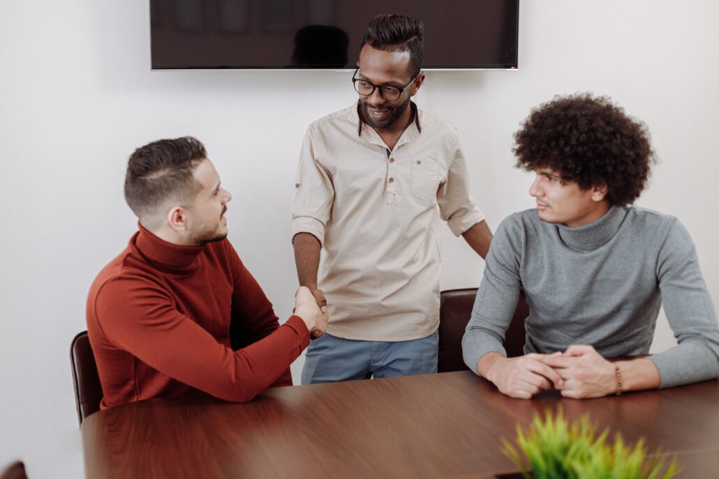 Professional coaching men in a collaborative workplace setting, with three colleagues engaged in discussion and a handshake during a team meeting.