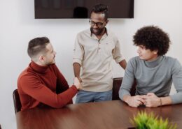 Professional coaching men in a collaborative workplace setting, with three colleagues engaged in discussion and a handshake during a team meeting.