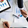 Two professionals shake hands over financial charts and business reports, illustrating how to start a life coaching business through planning, strategy, and partnership.