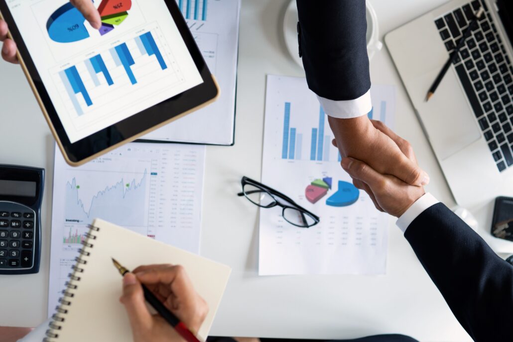 Two professionals shake hands over financial charts and business reports, illustrating how to start a life coaching business through planning, strategy, and partnership.