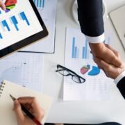Two professionals shake hands over financial charts and business reports, illustrating how to start a life coaching business through planning, strategy, and partnership.