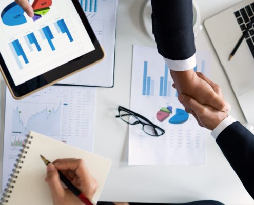 Two professionals shake hands over financial charts and business reports, illustrating how to start a life coaching business through planning, strategy, and partnership.